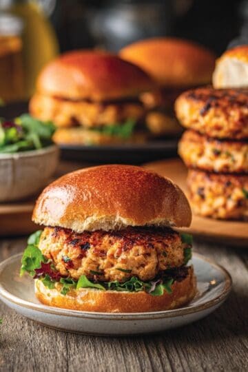 ground chicken burger on a small plate, wood table, platter of chicken patties in the background