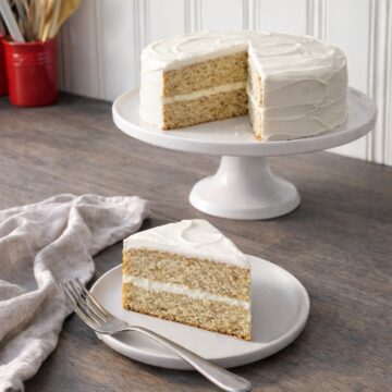 Bakery-style banana cake with cream cheese frosting, fully frosted with a middle layer, shown on a pedestal cake stand with a sliced piece plated in the foreground.