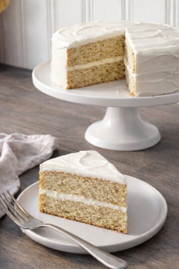 Bakery-style banana cake with cream cheese frosting, fully frosted with a middle layer, shown on a pedestal cake stand with a sliced piece plated in the foreground.
