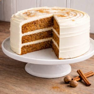Three-layer carrot cake with cream cheese frosting on a white pedestal stand, a slice removed to reveal even layers and moist crumb, styled with cinnamon sticks and whole nutmeg on a grey-stained oak butcher block countertop.