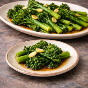Garlic broccolini served on a platter with a small plate in the foreground, photographed on a grey-stained wood kitchen counter.