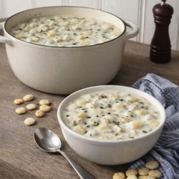 New England clam chowder served in a bowl with oyster crackers, shown on a wooden kitchen counter with an enamel cast iron pot in the background