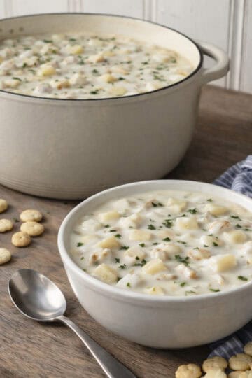 New England clam chowder served in a bowl with oyster crackers, shown on a wooden kitchen counter with an enamel cast iron pot in the background