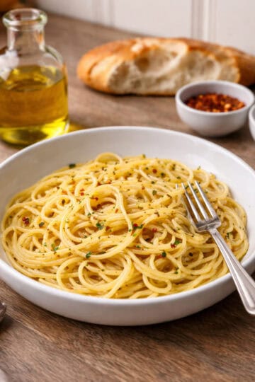 Spaghetti aglio e olio in a white bowl with olive oil bottle, chili flakes, finishing salt, torn loaf of bread, wine glass, and fork on a rustic wooden countertop
