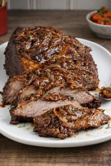 Beef Brisklet braised on a white platter, sliced showing tneder cernter on a wood counter top, small bowl of braised carrots in the backgound