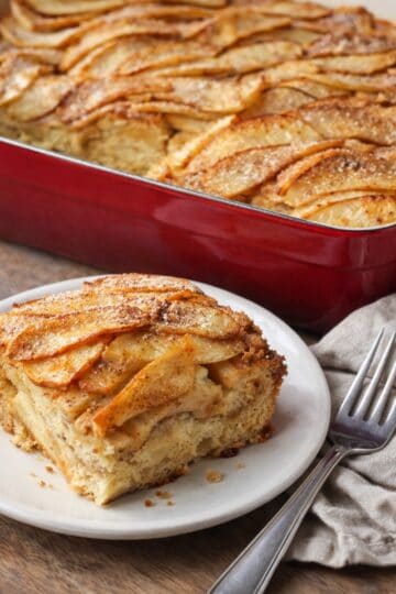 Apple French toast casserole in a red ceramic baking dish with thinly sliced apples on top, a corner slice removed, and a matching piece served on a white plate with a fork and rumpled napkin on a wood countertop.