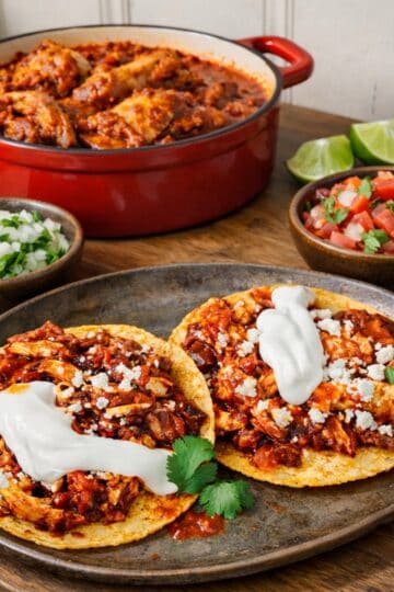 Chicken tinga tacos on a rustic platter with crema and cotija, set on a wood countertop with a red casserole of braised chicken and bowls of onion-cilantro relish and pico de gallo in the background