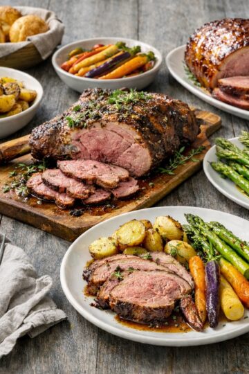 Easter dinner table with roasted leg of lamb, glazed ham, rainbow carrots, asparagus, and roasted potatoes on a grey wood table