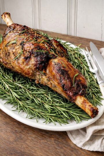 Roasted bone-in leg of lamb with rosemary on a white platter on a wooden countertop