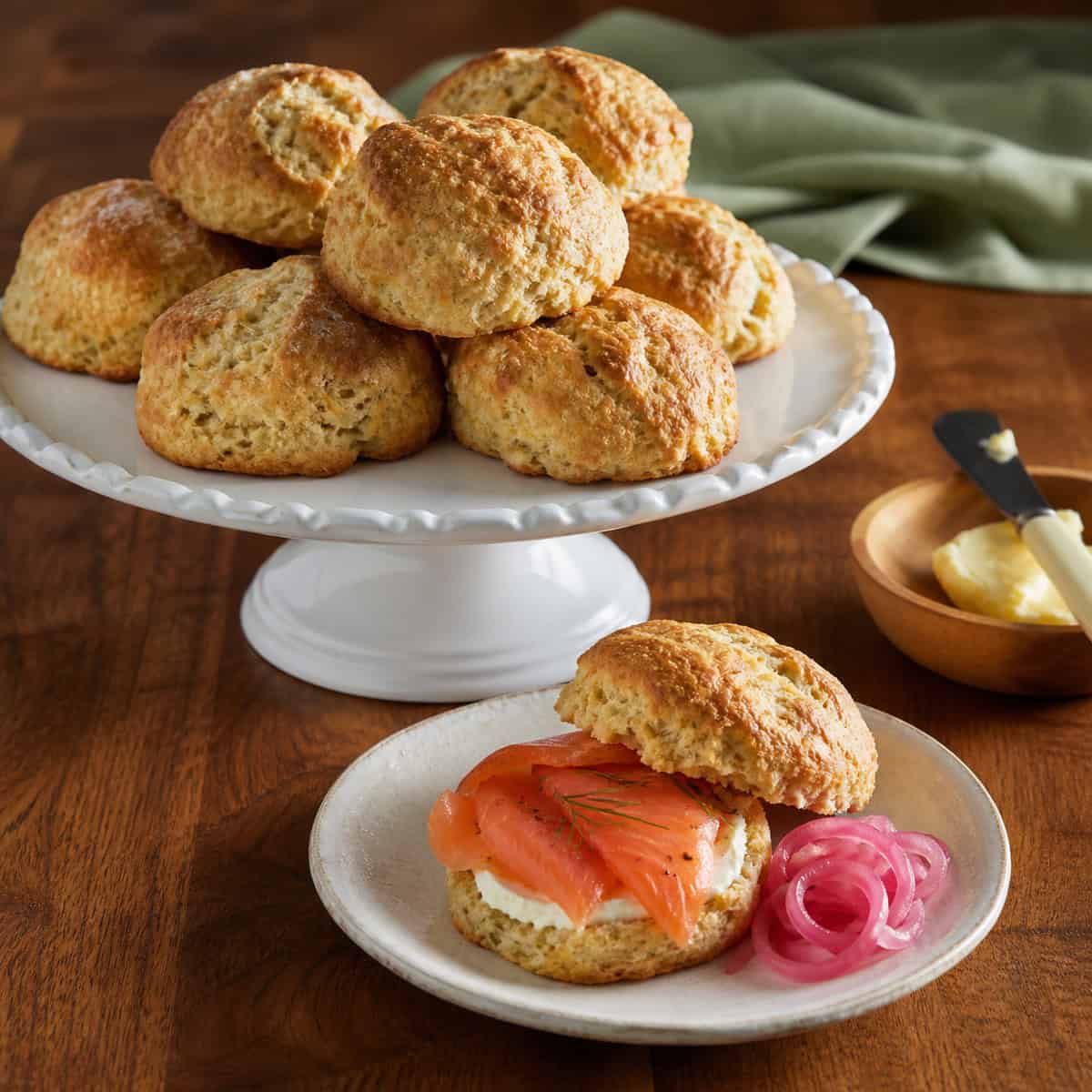 Irish brown soda scones on a white pedestal plate with a warm scone topped with smoked salmon and pickled onions on a small plate over a rustic wood countertop.